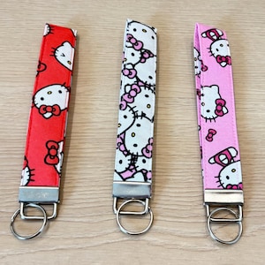 May include: Three fabric key fobs with metal hardware. The key fobs feature a repeating pattern of a cartoon cat with a bow. One is red, one is white, and one is pink. Each has a silver-colored key ring.