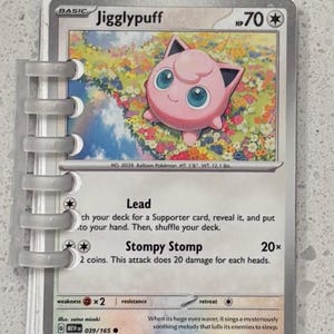 May include: A Jigglypuff Pokémon card, featuring a pink character with large blue eyes, set against a floral background. The card includes the text "Jigglypuff", "HP70", and attack descriptions. The card is held in a clear binder.