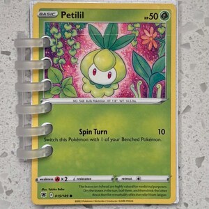 May include: A Petilil Pokémon trading card with a green and yellow design. The card features a cartoon image of a plant-like creature and the text "Petilil". The card is held in a clear, plastic binder.