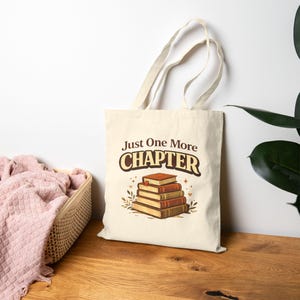 Könnte beinhalten: Cremefarbene Tragetasche mit dem Text "Just One More Chapter" in Braun und Weiß. Die Tasche zeigt einen Stapel Bücher in Braun- und Orangetönen mit dekorativen Elementen. Sie hat lange Henkel und ist aus einem leinwandartigen Material.