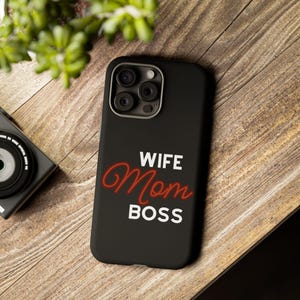 Könnte beinhalten: Schwarze Handyhülle mit den Worten "WIFE Mom BOSS" in Weiß und roter Schrift. Die Hülle hat einen Kameraausschnitt und liegt auf einer Holzoberfläche.