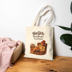 Könnte beinhalten: Cremefarbene Tragetasche mit dem Text "Hot Girls Read Books & Drink Matcha" in Braun. Die Grafik zeigt eine Person mit roten Haaren, die ein Buch liest und Matcha trinkt. Die Tasche hat zwei beige Griffe.