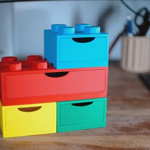 May include: A set of four brightly coloured storage drawers, designed to look like large toy building blocks. The drawers are in red, blue, yellow, and green. Each drawer has a curved handle. The drawers are stacked on a wooden surface.