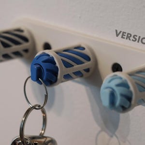 May include: A white key holder featuring three key storage units. Each unit has a blue interior and a white cage-like exterior. One unit holds a key ring with keys. The text "VERSION 2" is visible.