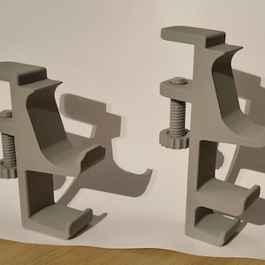 May include: Two grey plastic C-clamps. Each clamp features a screw mechanism for adjusting the grip. The clamps have a curved design and are made of a solid, matte grey plastic. They are designed to hold objects in place.