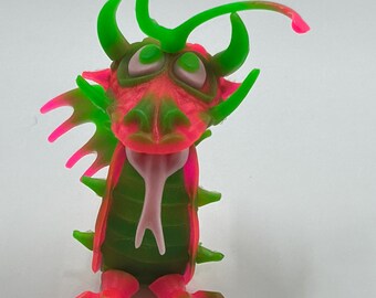May include: A colorful, whimsical dragon figurine. The dragon features a vibrant color scheme of green, pink, and red. It has horns, a spiky back, and large feet. The dragon's tongue is a light pink color.