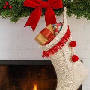 May include: A cream-colored knitted Christmas stocking with red fringe and pom-poms hangs from a fireplace. The stocking is filled with wrapped gifts. A red bow and garland with red berries are above the stocking.