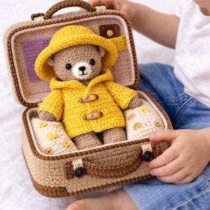 Può includere: Un orsacchiotto all'uncinetto con cappotto e cappello gialli, all'interno di una valigia abbinata. La valigia è beige con finiture marroni e un manico. L'orso è seduto su un cuscino con fiori gialli.