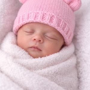 以下が含まれることがあります： 白い毛布に包まれた眠っている赤ちゃん。ピンク色のクマの耳が付いたニット帽をかぶっています。帽子と毛布が柔らかく居心地の良い雰囲気を作り出しています。赤ちゃんの顔が見え、目を閉じて穏やかな表情をしています。