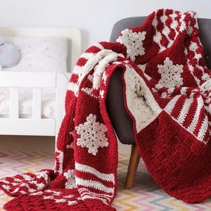 May include: A red and white crocheted blanket with snowflake and striped patterns. The blanket is draped over a gray chair in a room with a crib and patterned rug. The blanket's design features large white snowflakes.