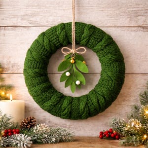 Könnte beinhalten: Ein grüner gestrickter Weihnachtskranz mit Mistelakzent, der vor einem Holzgrund hängt. Der Kranz ist mit einem dekorativen Mistelzweig mit goldenen und weißen Akzenten geschmückt, der an Naturfaden aufgehängt ist.