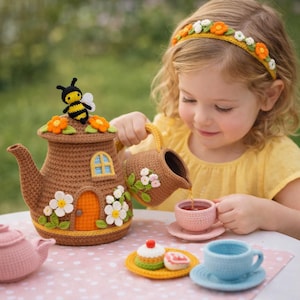 May include: A crocheted brown teapot with a house design, decorated with flowers and a bee, pours tea into a pink teacup. A matching creamer, teacup, and treats complete the tea party setting. A young girl wears a floral headband.