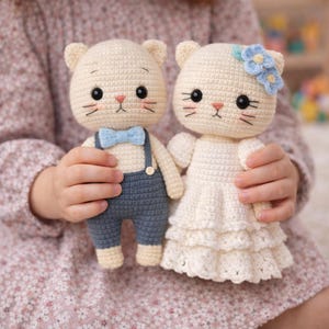 May include: Two handmade crochet cat dolls. One cat doll wears blue overalls and a blue bow tie. The other cat doll wears a white dress and a blue flower. Both dolls have black eyes, pink noses, and black whiskers. The dolls are held by a person.