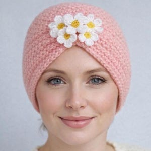 Puede incluir: Un turbante de punto rosa claro adornado con decoraciones en forma de margaritas blancas y amarillas. El sombrero está hecho de un material suave y texturizado y está diseñado para ser usado en la cabeza.