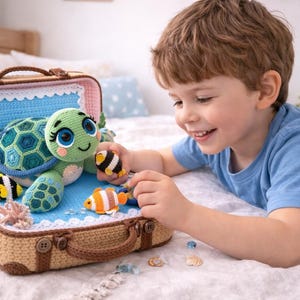Könnte beinhalten: Eine gehäkelte Schildkröte und Fischspielzeuge werden in einem kleinen Koffer ausgestellt. Die Schildkröte ist grün und blau, mit großen Augen. Der Koffer ist beige mit einem braunen Griff. Ein Kind lächelt und spielt mit den Spielzeugen.
