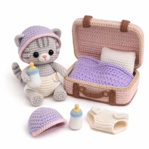 May include: A crocheted gray kitten doll wearing a lavender and pink hat and diaper, holding a bottle. A pink suitcase contains a blanket and pillow. Additional accessories include a hat, bottle, and diaper.