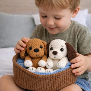 Pode incluir: Dois cachorros de pelúcia de crochê, um marrom e outro branco com orelhas marrons, sentados em uma cesta marrom com forro azul. Os cães têm olhos pretos e são feitos à mão. A cesta é feita de fio marrom.