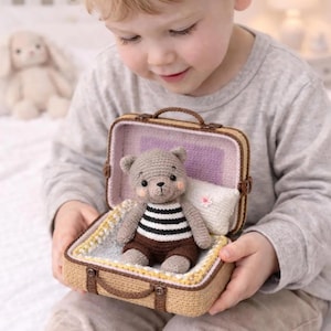 Puede incluir: Un oso de peluche de ganchillo con una camisa a rayas y pantalones cortos marrones está sentado dentro de una maleta en miniatura. La maleta es de color canela con un asa y ribetes marrones. También hay una pequeña almohada.