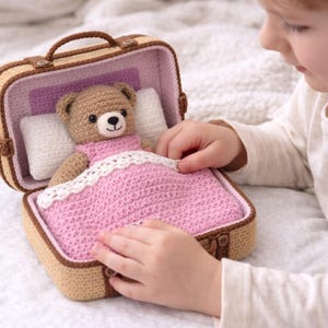 Può includere: Un orsacchiotto all'uncinetto in un letto a valigia in miniatura. L'orso indossa un vestito rosa ed è coperto da una coperta rosa bordata di pizzo bianco. La valigia è beige con finiture marroni e una maniglia. Mani di un bambino visibili.