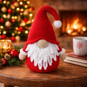 Pode incluir: Um gnomo de crochê vermelho e branco feito à mão, com um chapéu comprido e pontudo e uma barba branca. O gnomo tem um nariz e braços bege, e está sobre uma mesa de madeira. Uma árvore de Natal e uma lareira são visíveis.