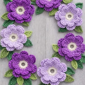 May include: A circular arrangement of seven crocheted flowers in shades of purple and lavender, each with a cream-coloured centre. Green crocheted leaves are positioned around each flower, creating a decorative floral design.