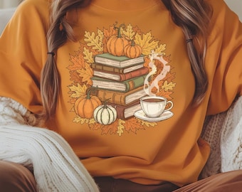 May include: Orange sweatshirt featuring a fall-themed graphic. The design includes a stack of books, pumpkins, autumn leaves, and a cup of coffee with steam rising. The overall aesthetic is cozy and autumnal.