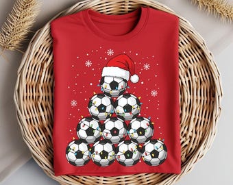 May include: Red t-shirt featuring a Christmas tree design made of soccer balls. The soccer balls are decorated with string lights and topped with a Santa hat. The shirt is folded in a wicker basket.