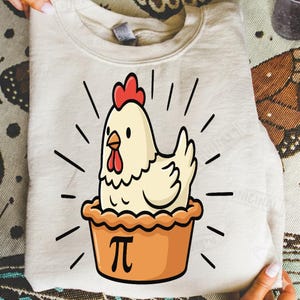 Pode incluir: Camisola creme com um desenho animado de uma galinha dentro de uma crosta de torta. A galinha tem uma crista e barbela vermelhas, com linhas pretas irradiando para fora. A crosta da torta é laranja com a letra grega pi.