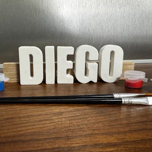 Könnte beinhalten: Der Name "DIEGO" in weißen 3D-Buchstaben auf einer Holzbasis. Kleine Töpfe mit blauer, roter und grüner Farbe sind zu sehen, zusammen mit zwei Pinseln, was auf eine kreative Aktivität hindeutet.