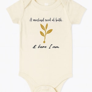 Könnte beinhalten: Cremefarbener Baby-Body mit kurzen Ärmeln. Auf der Vorderseite steht der Text "A mustard seed of faith.." über einem goldfarbenen Blattdesign und der Text "& here I am" darunter.
