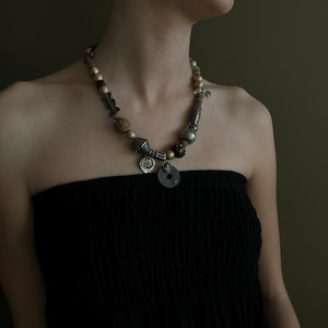 May include: A multi-strand necklace featuring a variety of beads in gold, silver, and brown tones. The necklace includes round, textured, and cylindrical beads, along with several metal pendants. The necklace is worn over a black strapless top.