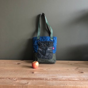 Könnte beinhalten: Eine blaue Jeans-Tragetasche mit einer Tasche aus Tarnstoff und grünen Riemen. Die Tasche steht auf einer Holzoberfläche mit einem roten Apfel davor.