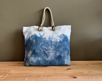 Puede incluir: Un bolso tote de lona con un estampado tie-dye azul. El bolso tiene asas gruesas de cuerda con detalles de cuero. El bolso está sobre una superficie de madera contra una pared de color neutro.