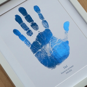 May include: A framed print with a blue handprint. The handprint has a metallic, textured finish. The print includes the text "Dad Forever in our hearts 1959-2022" in a simple font, set against a white background.