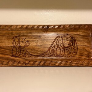 May include: A rectangular wooden tray with carved Arabic calligraphy. The tray has a decorative border and a rich, warm brown color. The calligraphy is also carved into the wood, creating a textured effect.