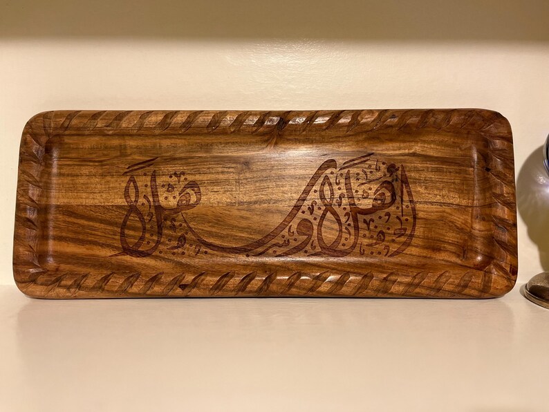 May include: A rectangular wooden tray with carved Arabic calligraphy. The tray has a decorative border and a rich, warm brown color. The calligraphy is also carved into the wood, creating a textured effect.