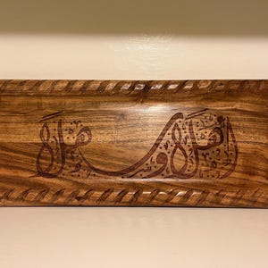 May include: Rectangular wooden tray with carved Arabic calligraphy. The tray has a dark brown finish with a decorative border. The calligraphy is also dark brown and is centered on the tray.