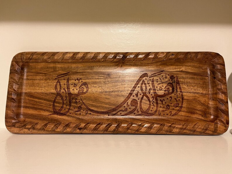 May include: Rectangular wooden tray with carved Arabic calligraphy. The tray has a dark brown finish with a decorative border. The calligraphy is also dark brown and is centered on the tray.