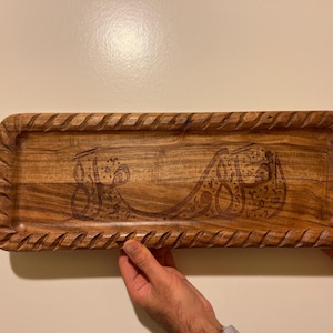 May include: A rectangular wooden tray with carved edges and an engraved Arabic script design. The tray is a warm brown color and appears to be handmade. The tray is being held by two hands.