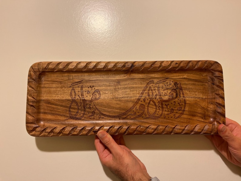 May include: A rectangular wooden tray with carved edges and an engraved Arabic script design. The tray is a warm brown color and appears to be handmade. The tray is being held by two hands.