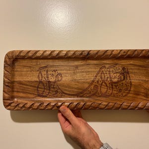 May include: A rectangular wooden tray with a carved, textured border. The tray features engraved Arabic calligraphy. The wood has a warm, natural tone. The tray is being held by two hands.