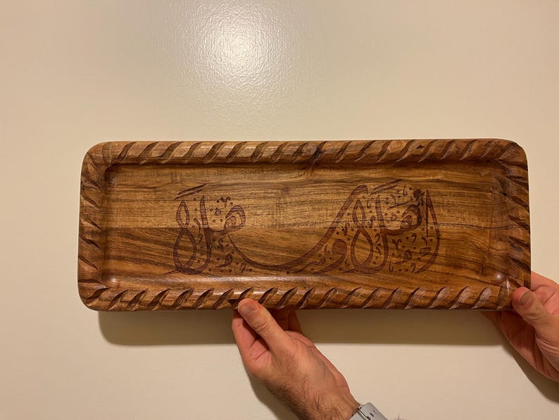 May include: A rectangular wooden tray with a carved, textured border. The tray features engraved Arabic calligraphy. The wood has a warm, natural tone. The tray is being held by two hands.