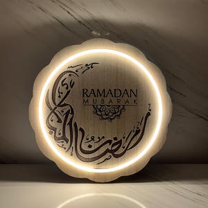 Op de afbeelding: Een ronde houten Ramadan Mubarak-bord met een witte LED-verlichting. Het bord toont Arabische kalligrafie en de Engelse tekst "Ramadan Mubarak". Het lichtbruine bord heeft een geschulpte rand.