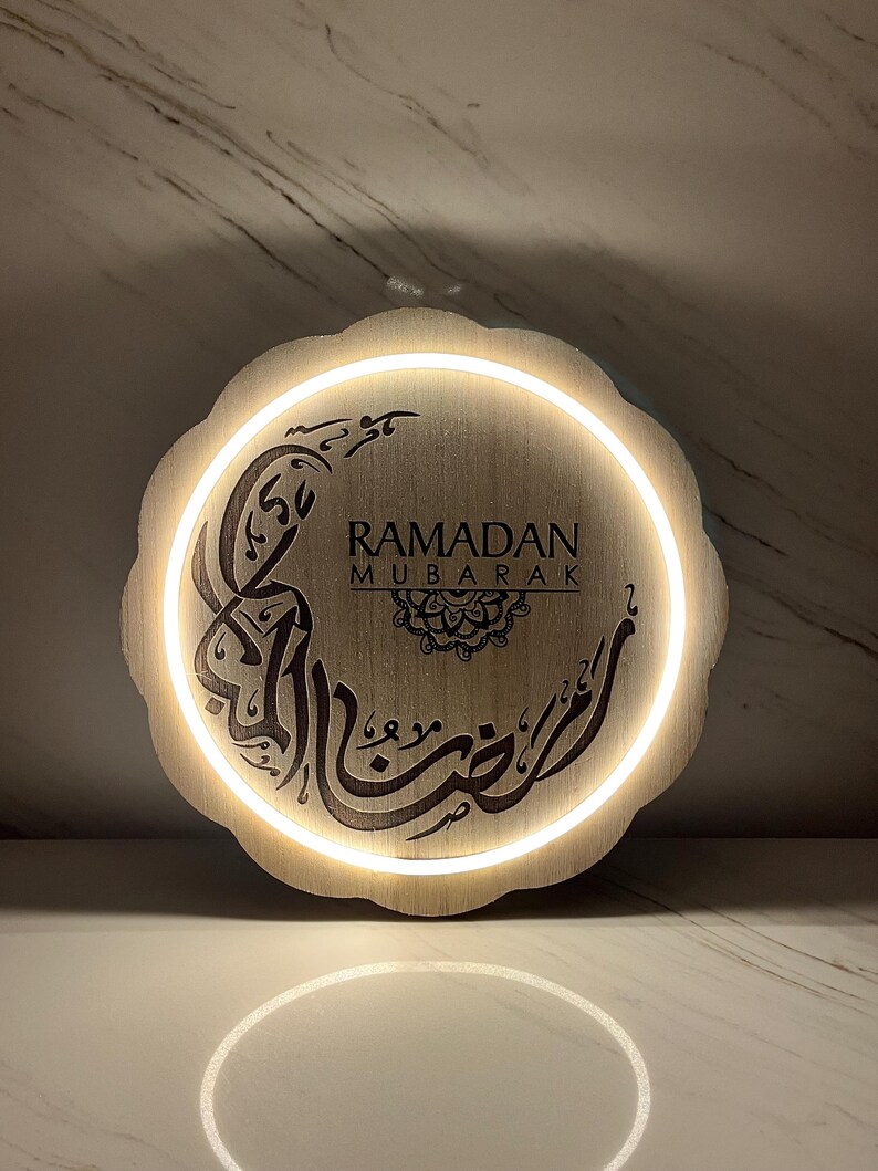 Op de afbeelding: Een ronde houten Ramadan Mubarak-bord met een witte LED-verlichting. Het bord toont Arabische kalligrafie en de Engelse tekst "Ramadan Mubarak". Het lichtbruine bord heeft een geschulpte rand.
