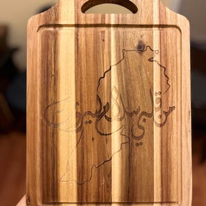 May include: Wooden cutting board with a handle and a carved design. The board features a map outline and Arabic script. The wood grain is visible, with a mix of light and dark brown tones. The board has a rectangular shape with a rounded top.
