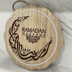 Op de afbeelding: Houten Ramadan Mubarak-bord met een geschulpte rand. Het bord heeft Arabische kalligrafie en de woorden "Ramadan Mubarak" in bruin tegen een lichte houtnerf achtergrond. Een wit cirkelvormig licht omringt de tekst.