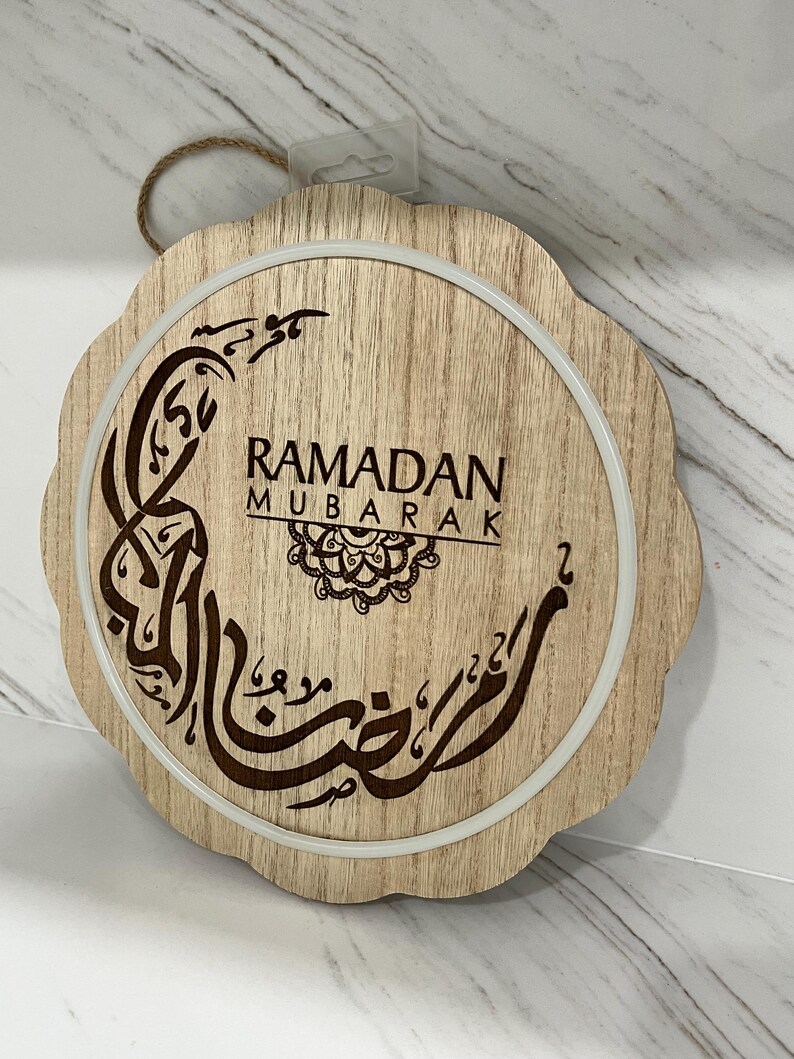 Op de afbeelding: Houten Ramadan Mubarak-bord met een geschulpte rand. Het bord heeft Arabische kalligrafie en de woorden "Ramadan Mubarak" in bruin tegen een lichte houtnerf achtergrond. Een wit cirkelvormig licht omringt de tekst.