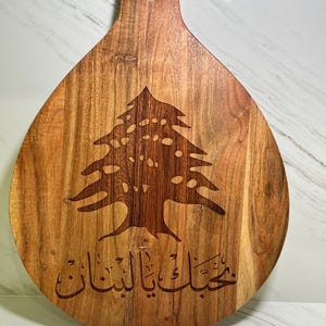 May include: A teardrop-shaped wooden serving board featuring a dark brown laser-engraved cedar tree emblem centered above Arabic script that reads "بحبك يا لبنان". The board has a smooth, polished finish with visible natural wood grain patterns and a circular handle hole at the top.