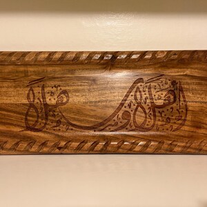 May include: Rectangular wooden tray with a carved design and a braided border. The tray is made of a light brown wood and features Arabic calligraphy in a darker brown color.