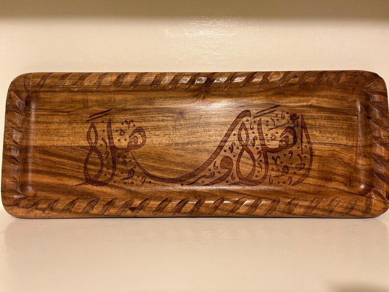 May include: Rectangular wooden tray with a carved design and a braided border. The tray is made of a light brown wood and features Arabic calligraphy in a darker brown color.